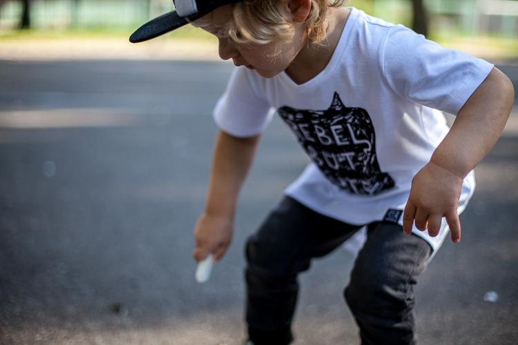 chłopiec w białej czapce z daszkiem kukukid i białej koszulce z napisem rebel but cute bawi się na asfaltowym boisku