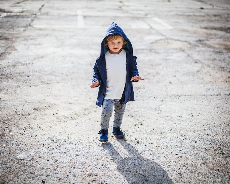 chłopiec w niebieskiej bluzie z kapturem, zamszowych bucikach i szarych legginsach spaceruje po parkingu