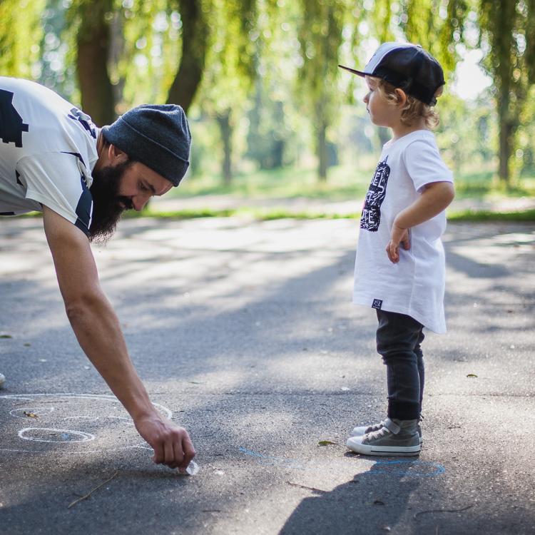 tata z brodą ubrany w białą koszulkę w czarny print wearso organic bawi się z synem ba asfaltowym boisku