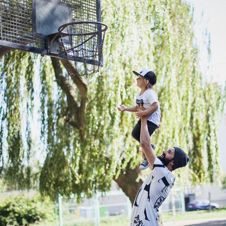 brodaty tata w koszulce wear so organic i czapce kukukid bawi się z synem na boisku koszykówki