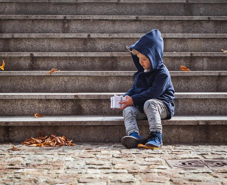 chłopiec w zamszowych bucikach i szarych legginsachi bawełnianym płaszczyku z kapturem sziedzi na kamiennych schodach
