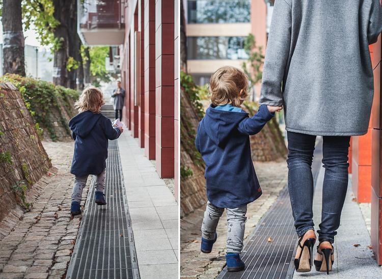 mama w szarym wełnianym płaszczu i czarnych rurkach spaceruje z malutkim synkiem po chodniku