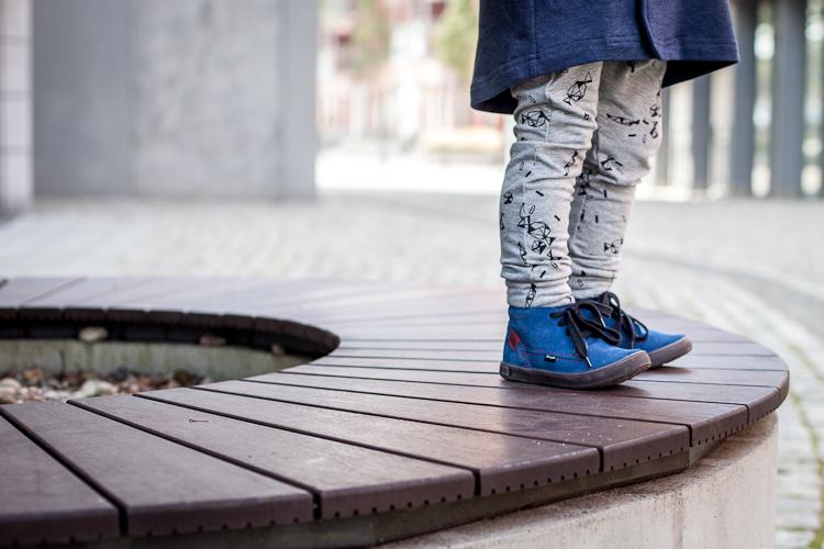chłopiec w niebieskich zamszowych bucikach i szarych legginsach w czarny print stoi na ławce