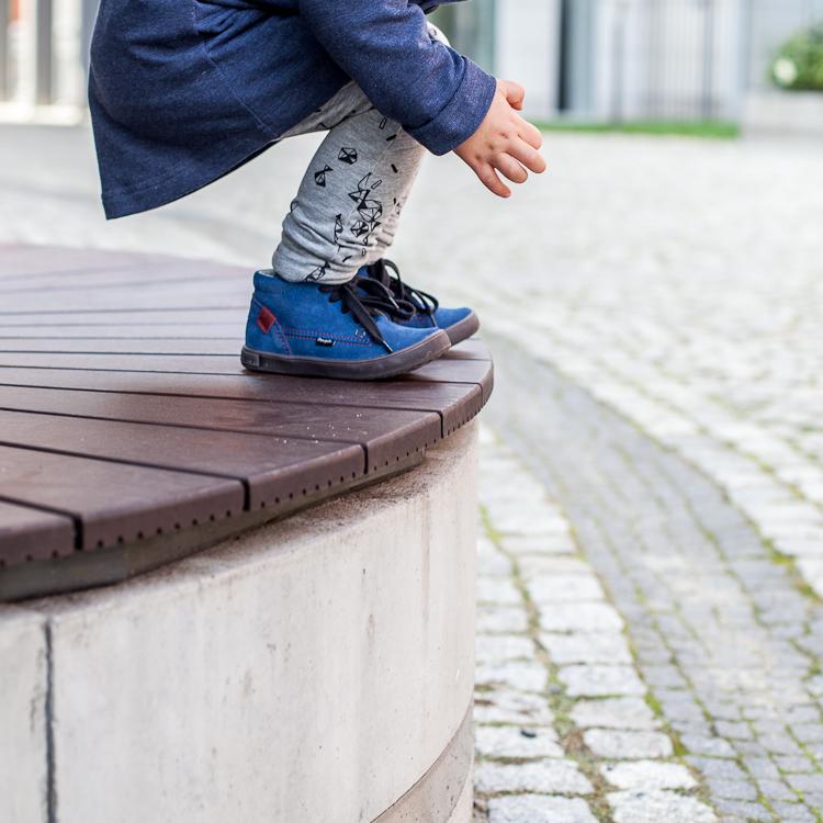 chłopiec w niebieskich zamszowych bucikach i szarych legginsach w czarny print skacze po ławce