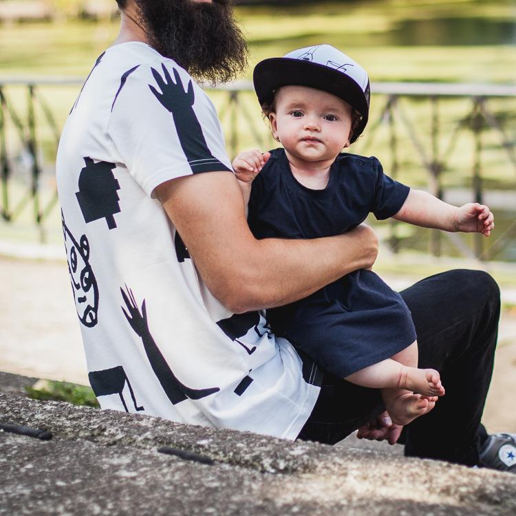 brodaty tata trzyma na rękach małą córeszkę, stojąc na boisku ubrany w koszulkę w czarny print i czapkę z daskiem