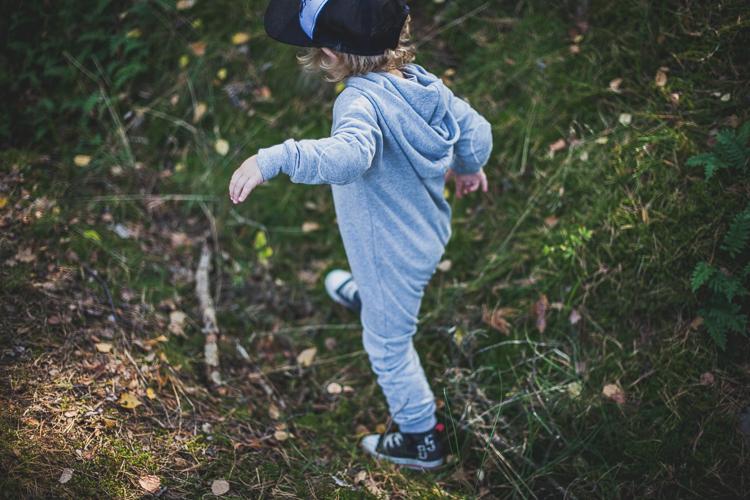 chłopiec ubrany w szary rampers i czapkę z daszkiem spaceruje po mchu na leśnej polanie