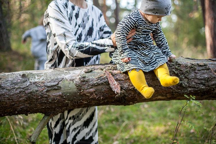 mama w czarno białej sukience Mainio sadza malutką córeczkę w żółtych podkolanówkach z błyskawicami na pniu drzewa