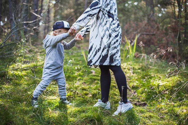 chłopiec ubrany w szary rampers radośnie podskakuje ze swoją mamą na leśnej ścieżce